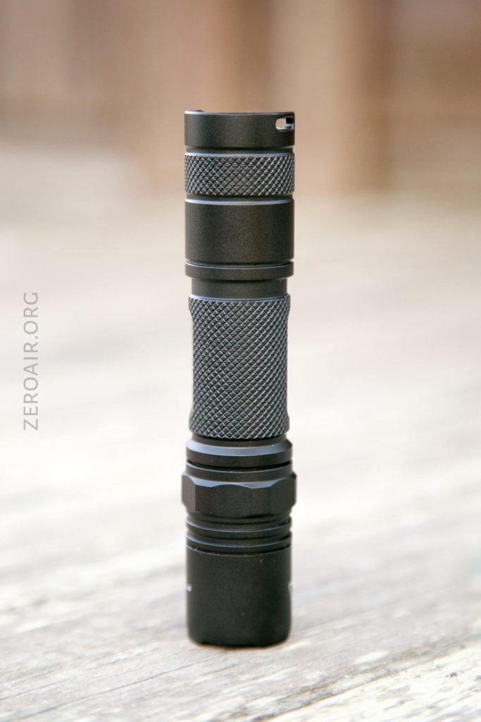 A black, compact, cylindrical flashlight stands upright on a light wooden surface, with textured grip sections and a flat top. The background is blurred, and the word ZEROAIR.ORG is faintly visible along the left side.