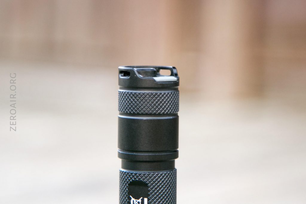 A close-up of the top section of a black, textured flashlight, showing a flat, notched metal bezel against a blurred neutral background. The word ZEROAIR.ORG is faintly visible on the left side.