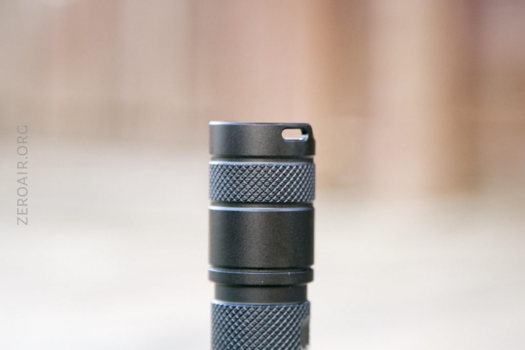 A close-up of a compact, black, cylindrical flashlight standing upright on a light surface with a blurred background. The side of the flashlight features textured grip sections. The word ZEROAIR.ORG is faintly visible on the left.