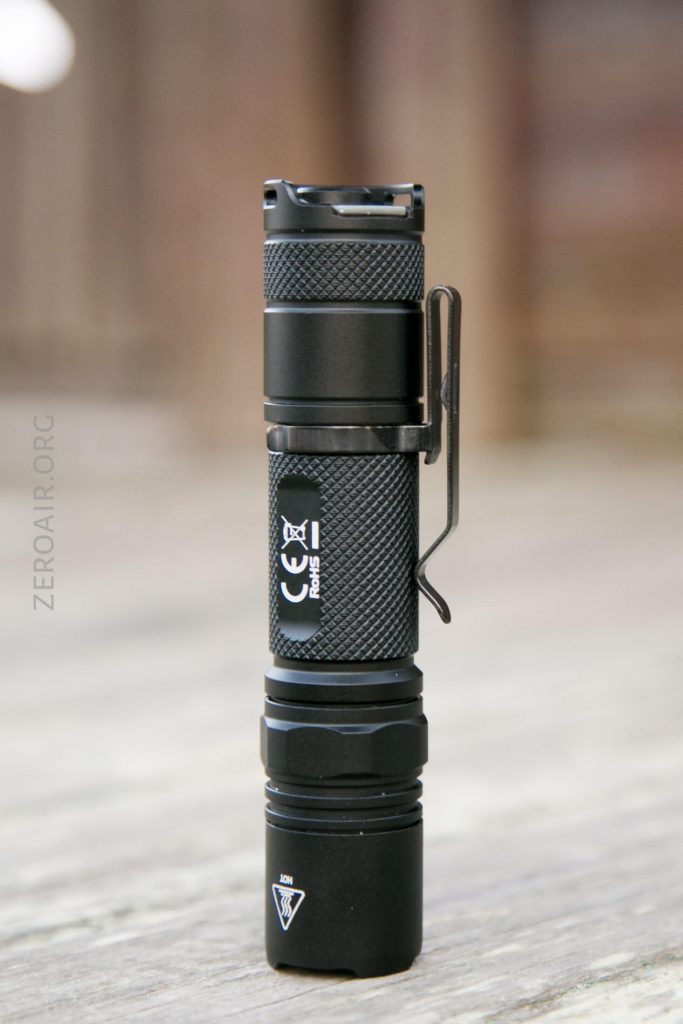 A black, compact flashlight with a textured grip, metal pocket clip, and visible regulatory markings stands upright on a wooden surface.