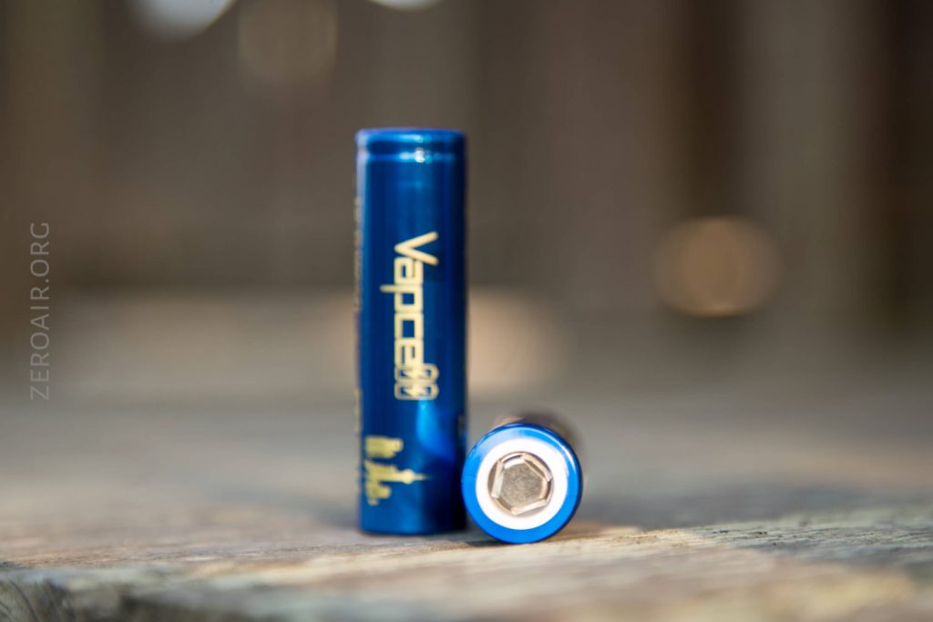 A blue cylindrical Vapcell battery stands upright next to another identical battery lying horizontally with its terminal facing forward, both on a wooden surface. The background is blurred.