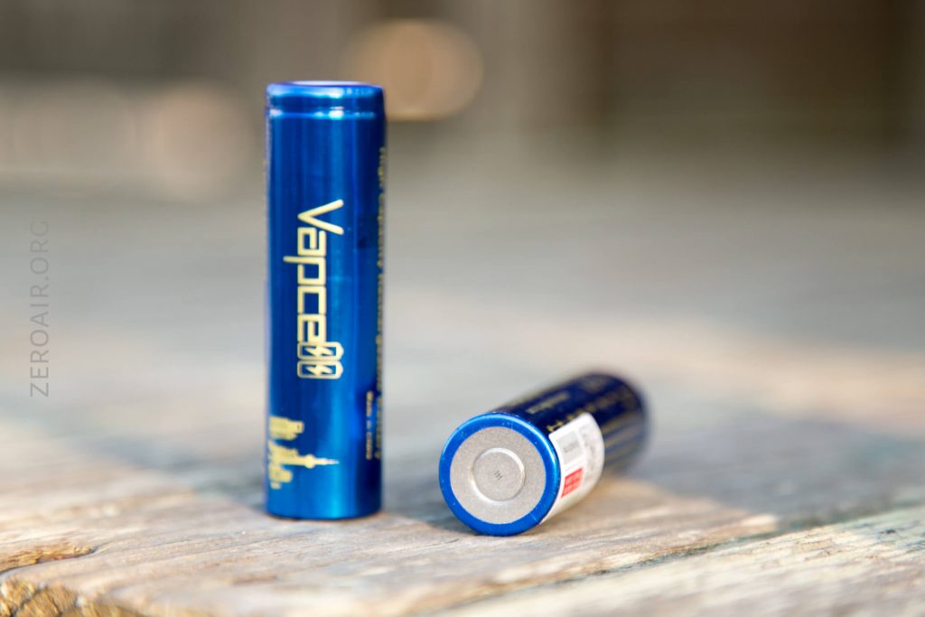 Two blue cylindrical batteries labeled Vapcell rest on a wooden surface; one is standing upright, and the other is lying down with the flat terminal side visible. The background is blurred.