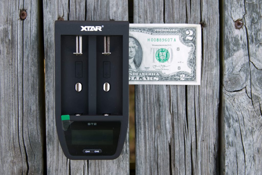 A black XTAR battery charger with two slots is placed on a wooden surface next to a US two-dollar bill. The items are arranged parallel, and the charger has a digital display at the bottom.