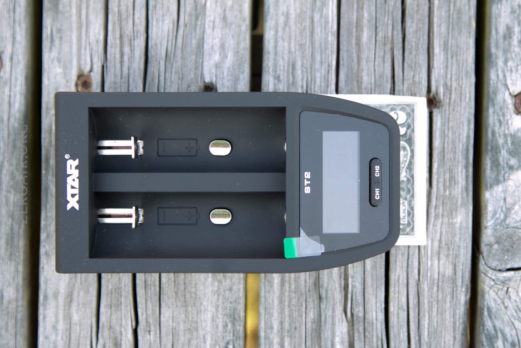 A black XTAR ST2 battery charger with two slots is placed on a weathered wooden surface. A partially visible playing card is underneath the right side of the charger.