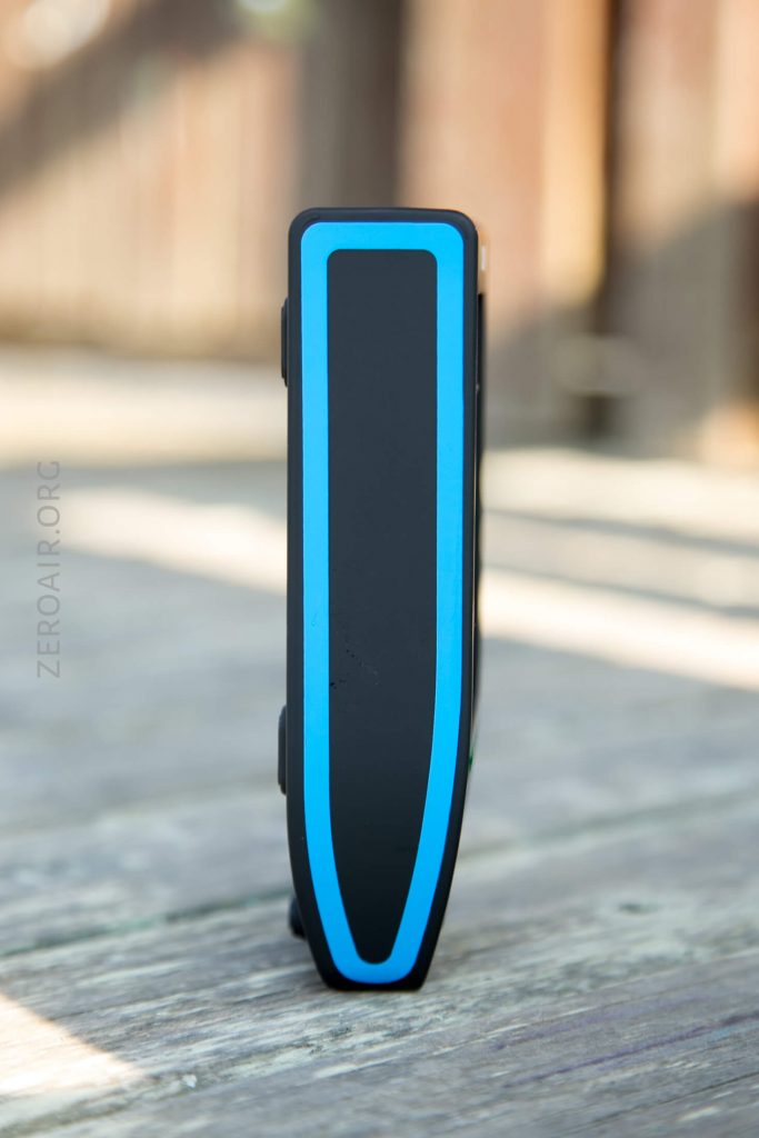 A black rectangular electronic device with a blue outline stands upright on a wooden surface, shown from the front. The background is blurred.