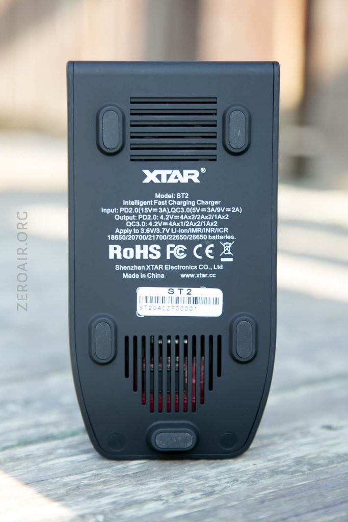 Close-up of the bottom of an XTAR ST2 battery charger, showing product information, safety certifications, input and output specifications, company name, and ventilation slots. Background is blurred wood surface.