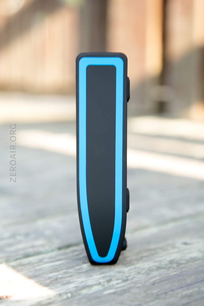 A small black rectangular electronic device with a blue border is standing upright on a wooden surface. The background is blurred.