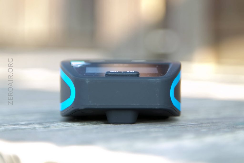 A small, rectangular electronic device with a screen and blue accents sits on a wooden surface. The side facing the camera shows a protruding button and the text ZEROAIR.ORG is visible on the background.