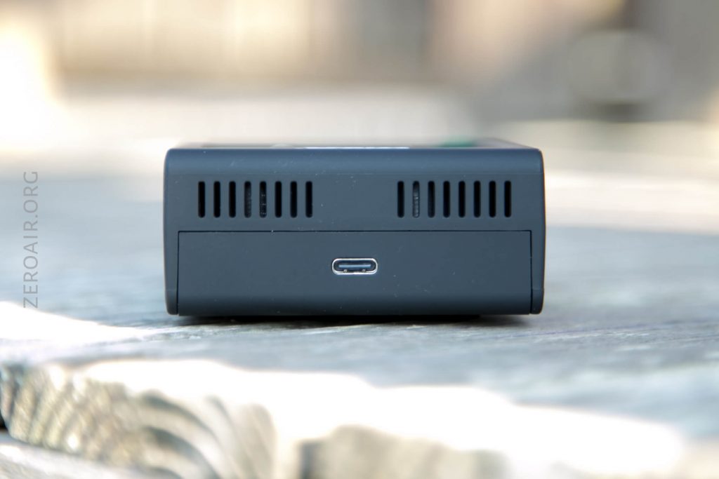 A close-up of a small black electronic device showing its back side, which features ventilation slots and a single USB-C port, placed on a wooden surface.