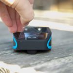 A hand is peeling a clear protective film off the screen of a small black electronic device with blue accents, which is resting on a wooden surface.