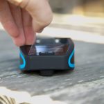 A hand is peeling a protective film off the screen of a small black device with blue accents, resting on a weathered wooden surface.