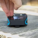 A hand is adjusting a small black and blue digital device with a screen and buttons, placed on a wooden surface. The device is photographed at close range in natural light.
