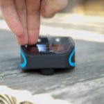 A close-up of a persons fingers pressing down on a small, black and blue electronic device placed on a wooden surface. The device has a rectangular screen and two visible buttons.