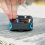 A hand holding a small plastic bag is placing or picking up an SD card from a black and blue electronic device sitting on a wooden surface.