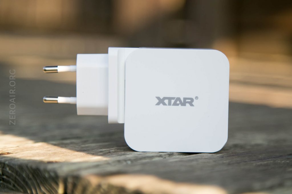 A white XTAR-branded wall charger with two round prongs is shown resting on a wooden surface. The logo XTAR is displayed on the side of the charger.