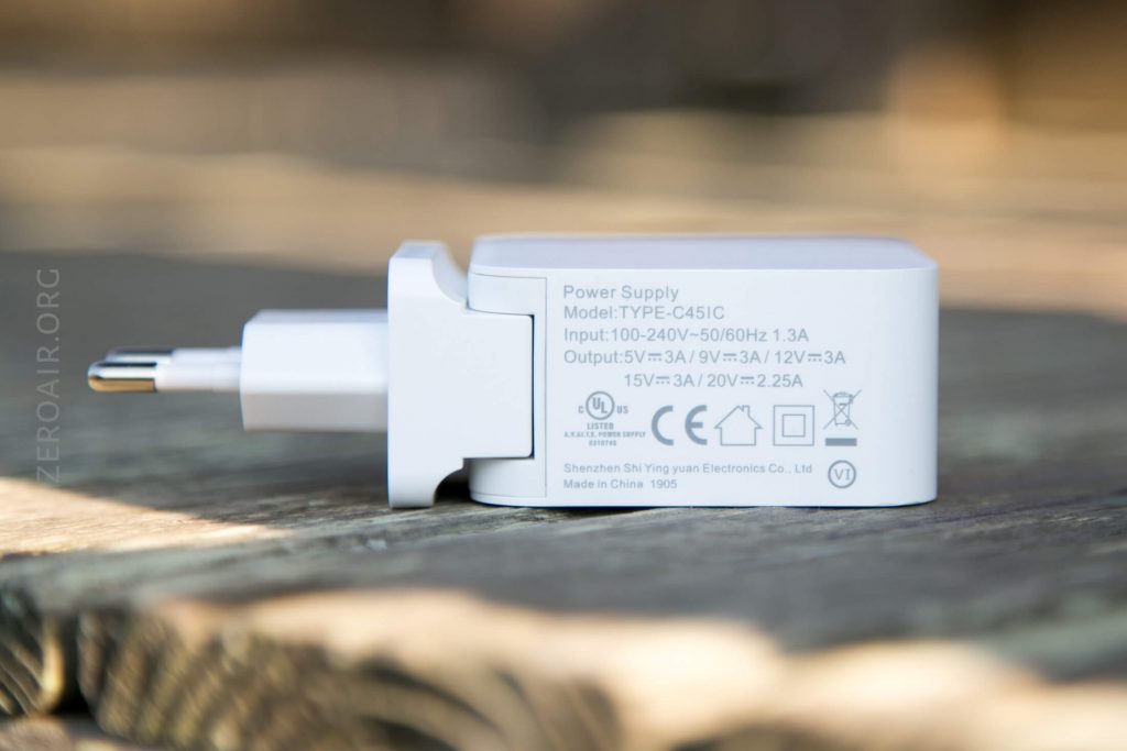 A white power adapter with prongs and technical specifications printed on the side, including voltage, current, and manufacturer information. The adapter is placed on a wooden surface with a blurred background.