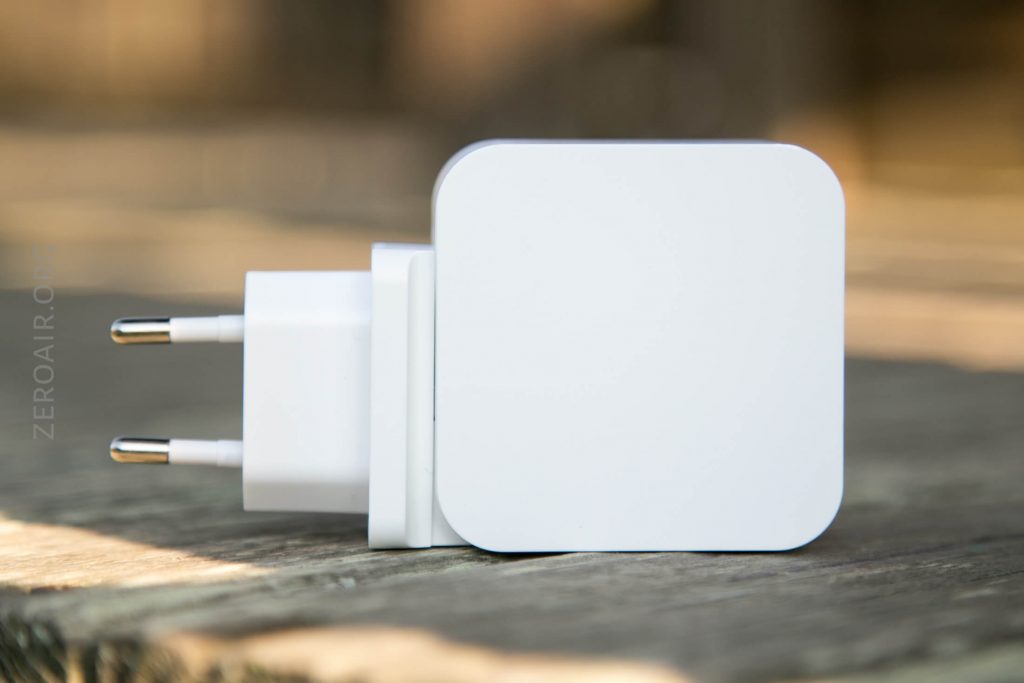A white rectangular wall charger with two round prongs lies on its side on a wooden surface, with a blurred background.