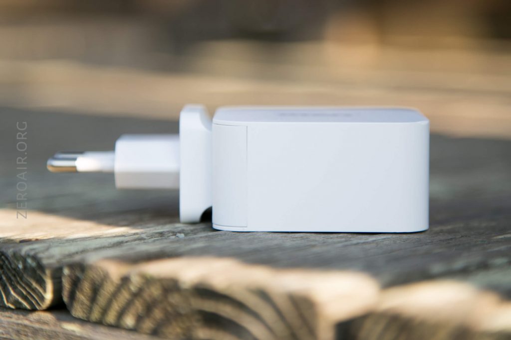 A white wall charger with a European plug is lying on its side on a wooden surface. The background is blurred, and the charger is photographed in natural light.