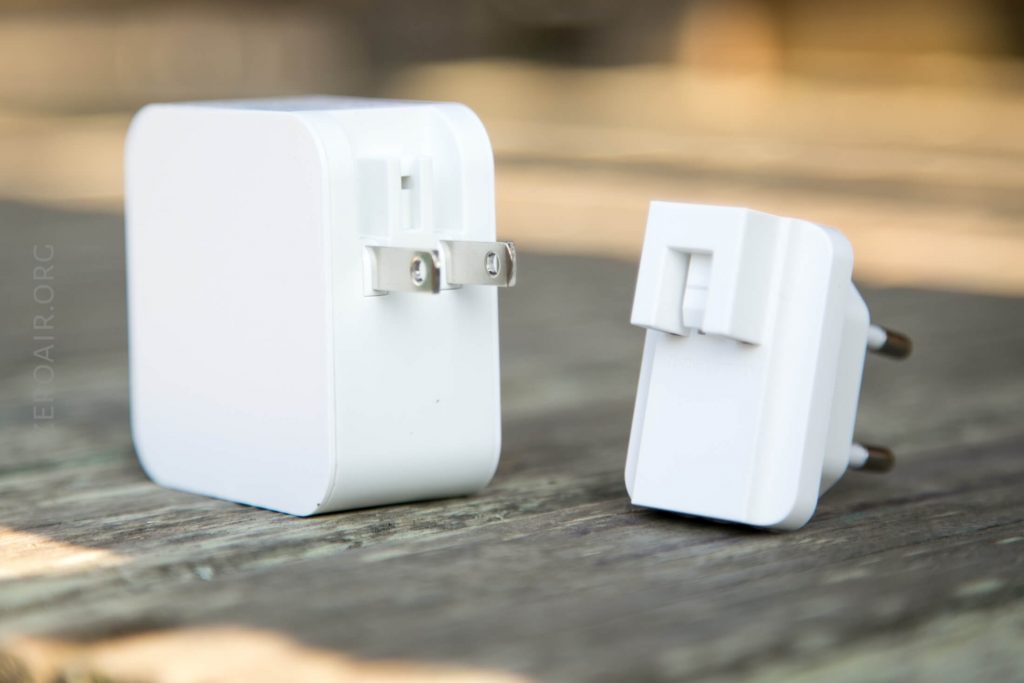 Two white electrical adapters are placed on a wooden surface. One adapter has flat prongs, while the other has round prongs. Both adapters are standing upright and shown from slightly different angles.