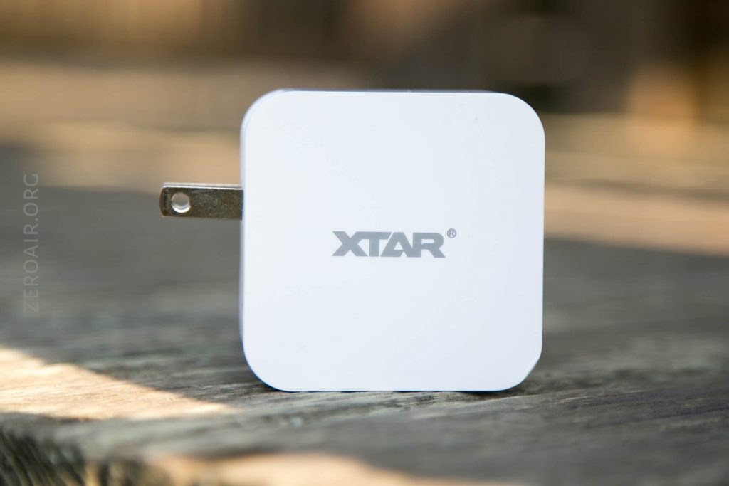 A white, square-shaped XTAR wall charger with a single metal plug prong is placed upright on a wooden surface. The background is blurred and out of focus.