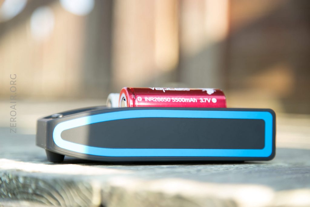 A black device with a blue stripe is placed on a wooden surface with two red 18650 batteries on top, one labeled INR 26650 5500mAh 3.7V. The background is blurred.