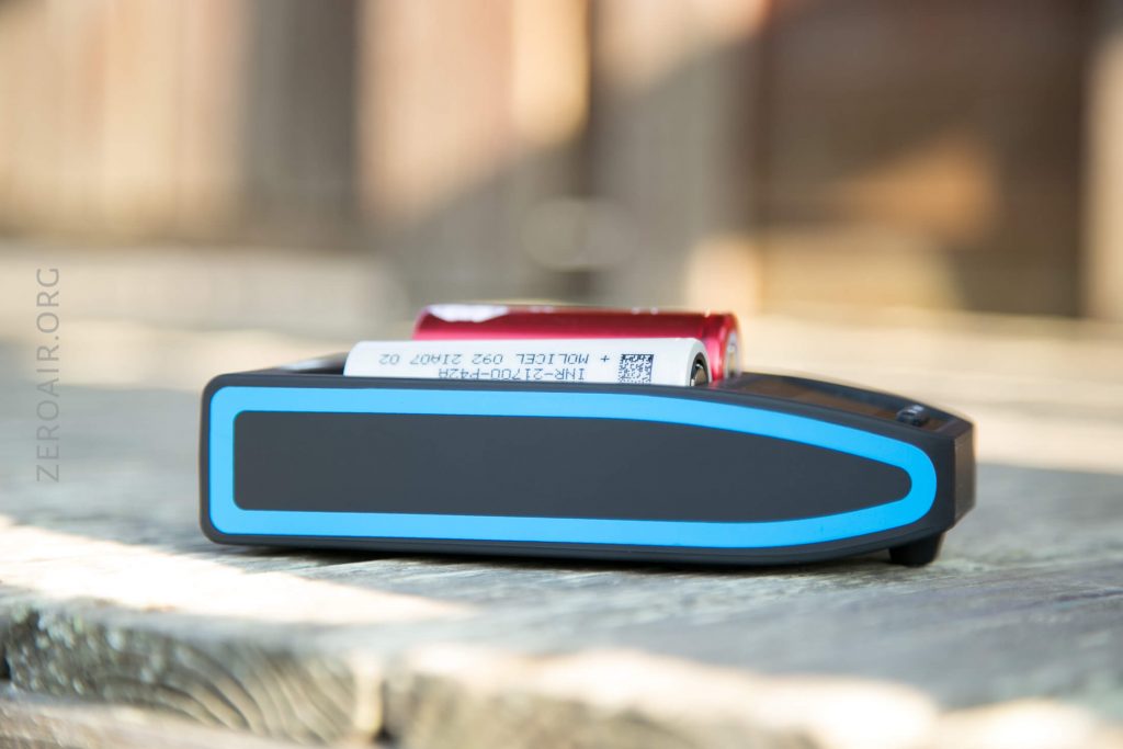 A rectangular battery charger with a blue trim sits on a wooden surface outdoors with a cylindrical red battery placed on top of it.
