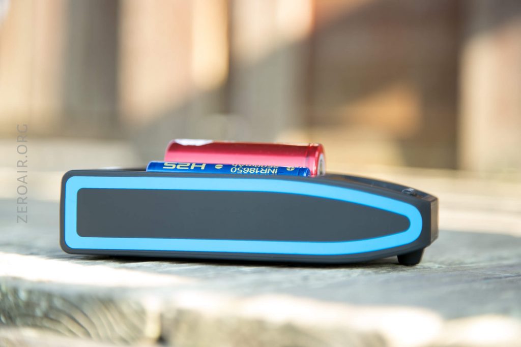 A black and blue battery charger with an 18650 lithium-ion battery inserted, resting on a wooden surface with a blurred background.