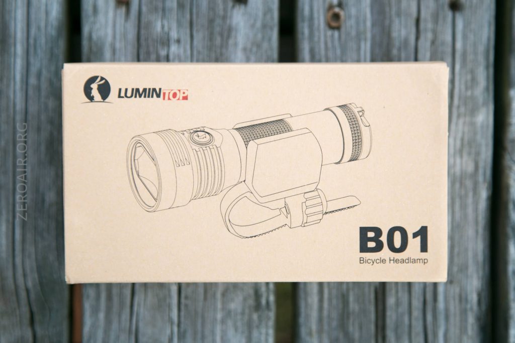 A cardboard box labeled LUMINTOP B01 Bicycle Headlamp shows a line drawing of a headlamp mounted on a bicycle handlebar, placed on a weathered wooden surface.