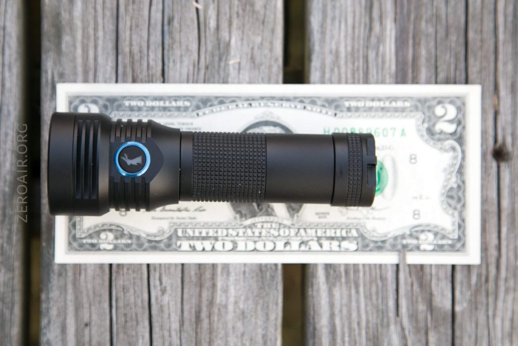 A black flashlight with a textured grip is placed horizontally on top of a U.S. two-dollar bill, both resting on a weathered wooden surface.