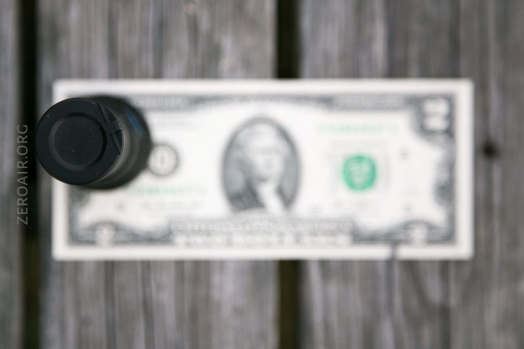 A black cylindrical object stands upright on a wooden surface, with a blurred U.S. one-dollar bill placed horizontally underneath it. The background appears out of focus.