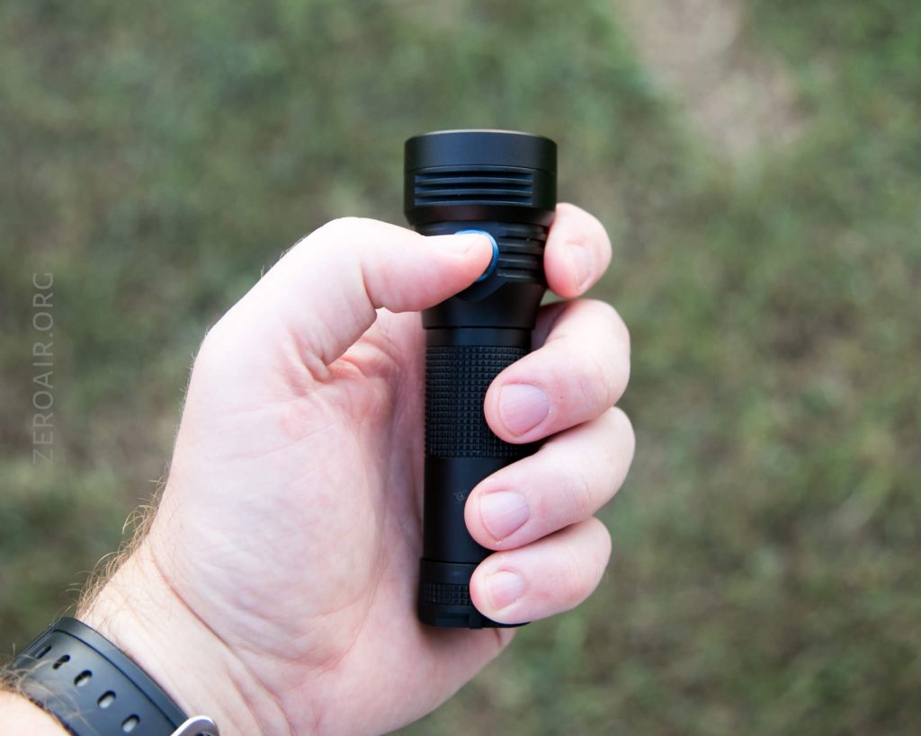 A person with a light skin tone holds a black flashlight in their right hand, thumb on the side button. The person is wearing a dark wristwatch. The background is grassy and out of focus.