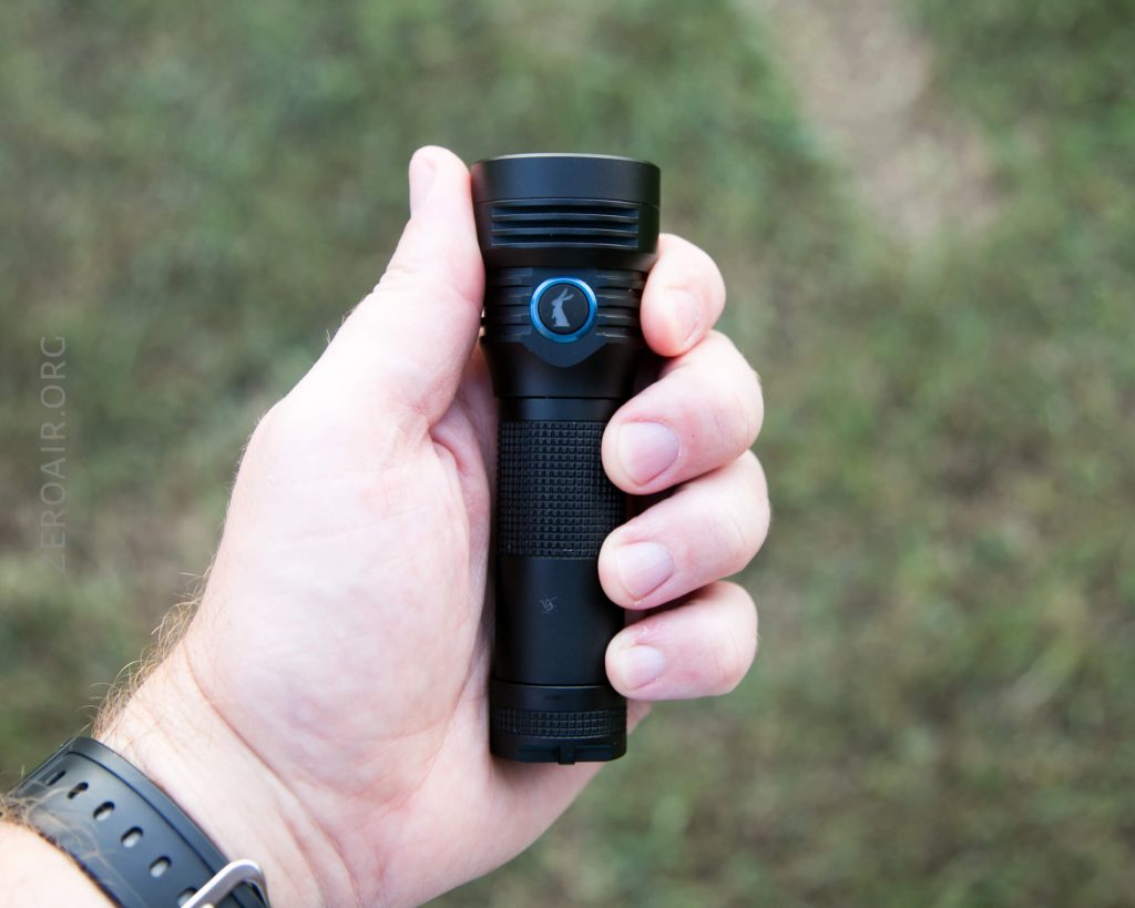 A hand holding a black flashlight with a blue power button, against a blurred grassy background. The person is wearing a gray wristwatch.