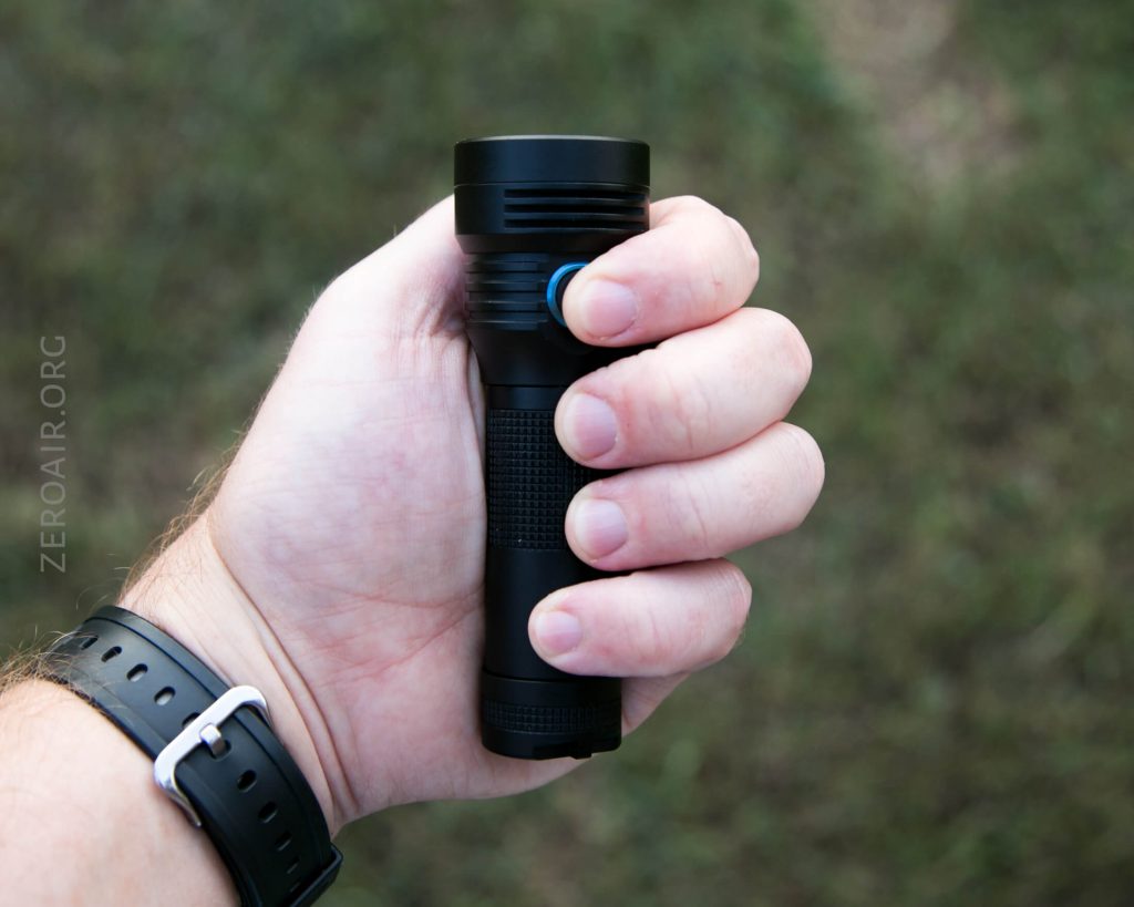 A person’s hand holding a black flashlight with a blue button, outdoors on a grassy background. The person is wearing a black wristwatch with a silver buckle.