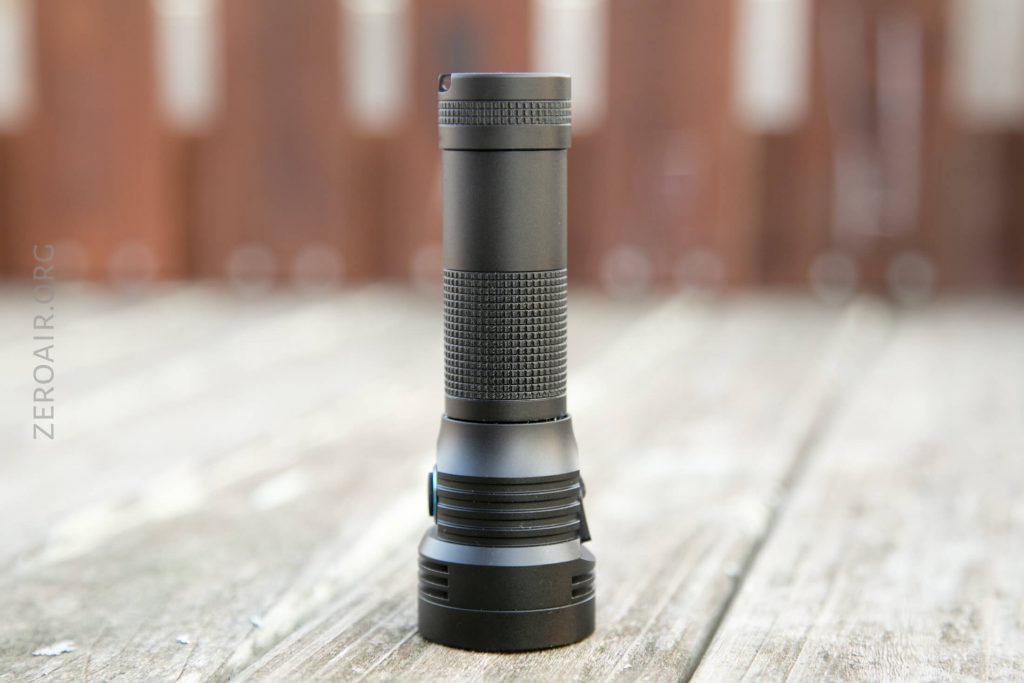 A black cylindrical flashlight with a textured grip stands upright on a wooden surface, with a blurred brown and white background.