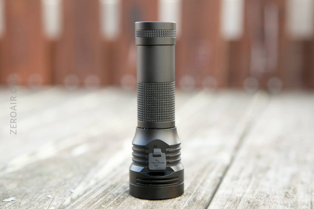 A black cylindrical flashlight stands upright on a wooden surface, with textured grip and a clip on the body. The background is blurred with brown vertical lines.
