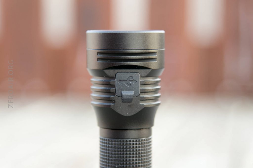 A close-up view of a black flashlight standing upright, showing a USB charging port symbol on the head. The background is out of focus with warm brown tones.