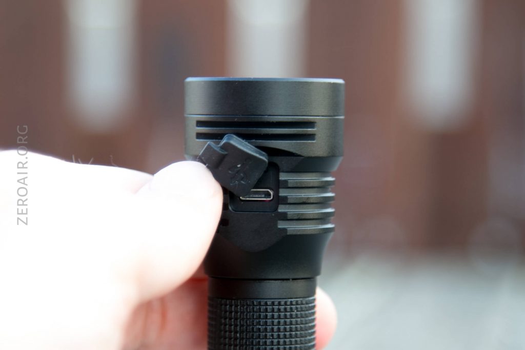 A hand lifts the rubber cover of a black flashlight to reveal a USB charging port. The background is blurred. The words ZEROAIR.ORG are visible along the left edge of the image.