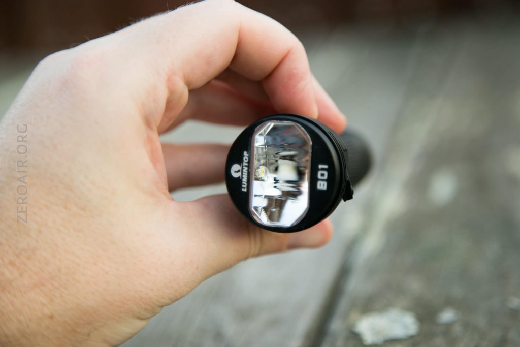 A hand holds a black flashlight outdoors, with the lens facing the camera. The flashlight has the model number B01 and LUMINTOP branding visible on its head. The background is a weathered wooden surface.
