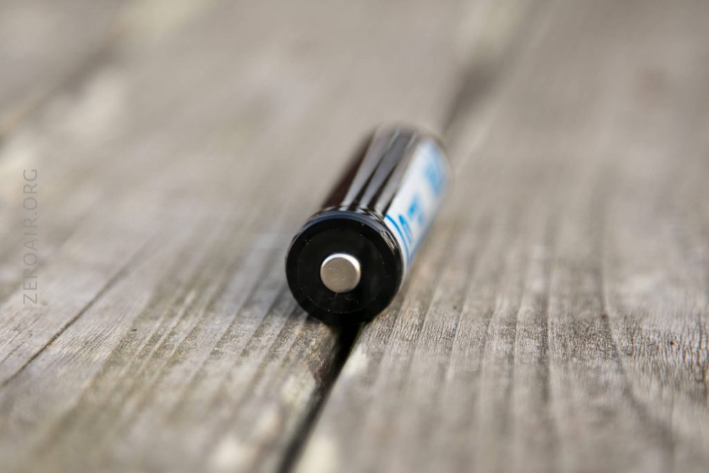 A close-up of a cylindrical AA battery lying horizontally on a weathered wooden surface, with the positive terminal facing the camera.