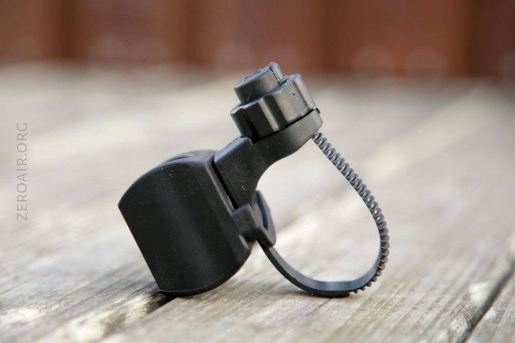 A black plastic bike accessory with an adjustable strap and dial, resting on a wooden surface. The device appears to be a mounting bracket, possibly for attaching a light or similar object to a bicycle.
