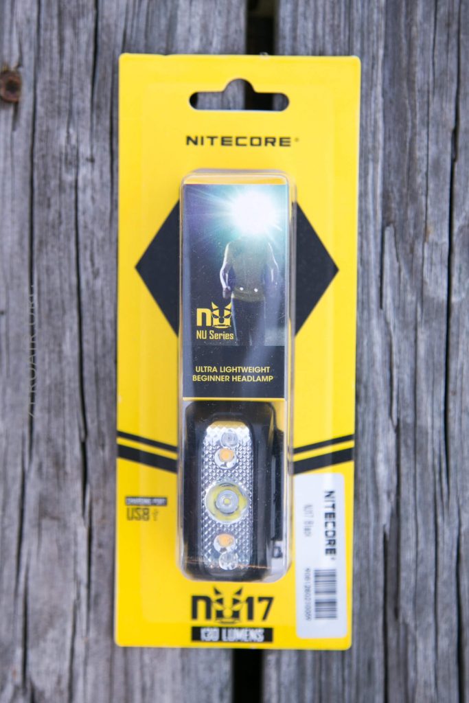 A Nitecore NU17 headlamp in its original yellow and black packaging, labeled Ultra Lightweight Beginner Headlamp, is placed on a weathered wooden surface. The package shows USB compatibility and 130 lumens brightness.