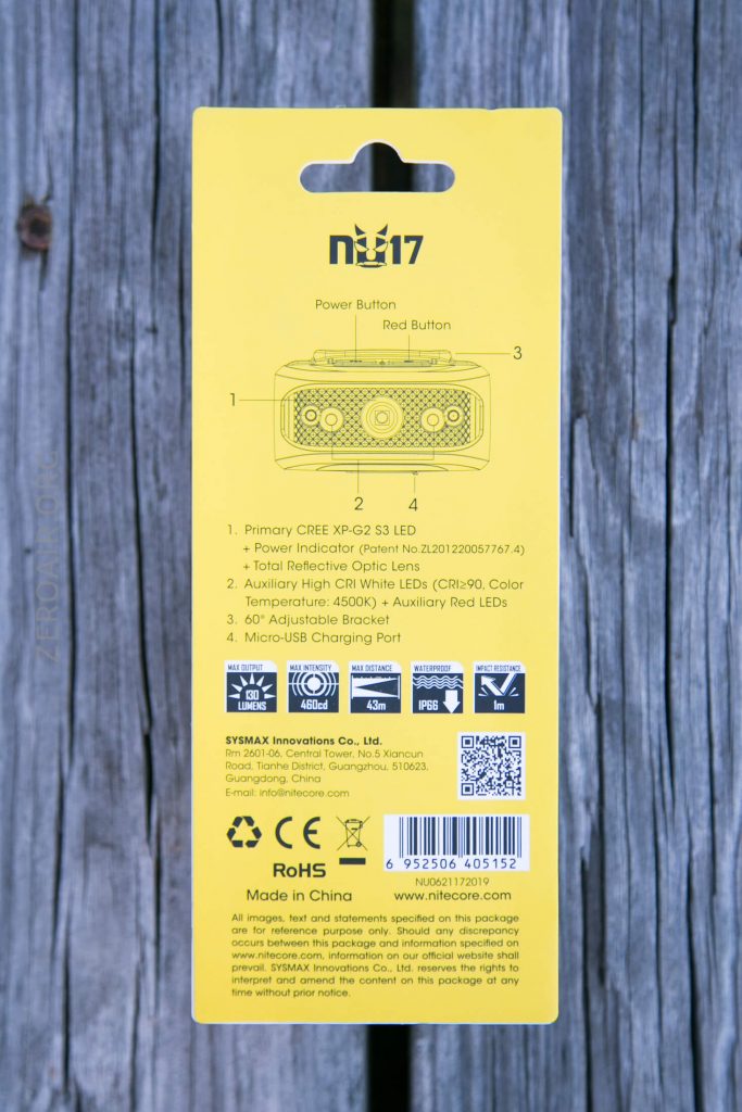 The back of a Nitecore NU17 headlamp package displays labeled product features, diagrams, a barcode, certification marks, manufacturer information, and a yellow background, all placed on a wooden surface.