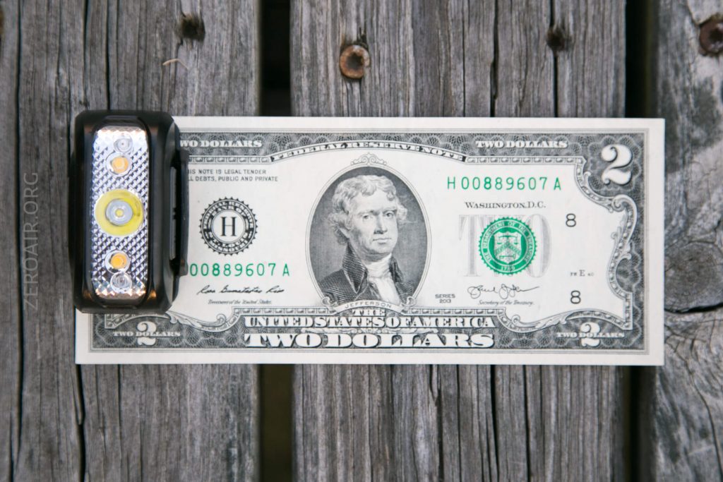 A small flashlight is placed next to a two-dollar bill on a wooden surface, showing the size comparison between the objects.