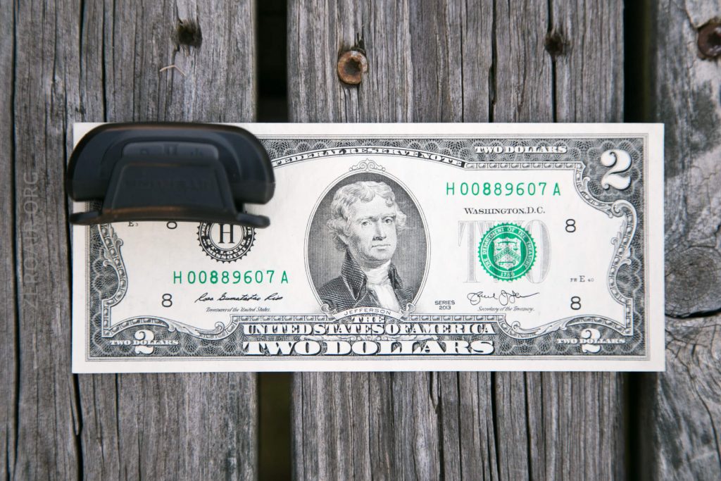 A U.S. two-dollar bill is secured to a weathered wooden surface with a black plastic clip, partially covering the left side of the bill featuring Thomas Jefferson’s portrait.