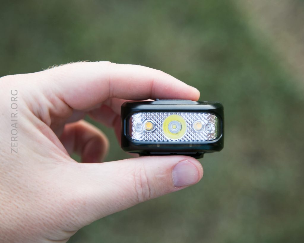 A hand holding a small black LED headlamp with three visible LED bulbs on the front, against a blurred outdoor background. The text ZERO-AIR.ORG is faintly visible along the left side.