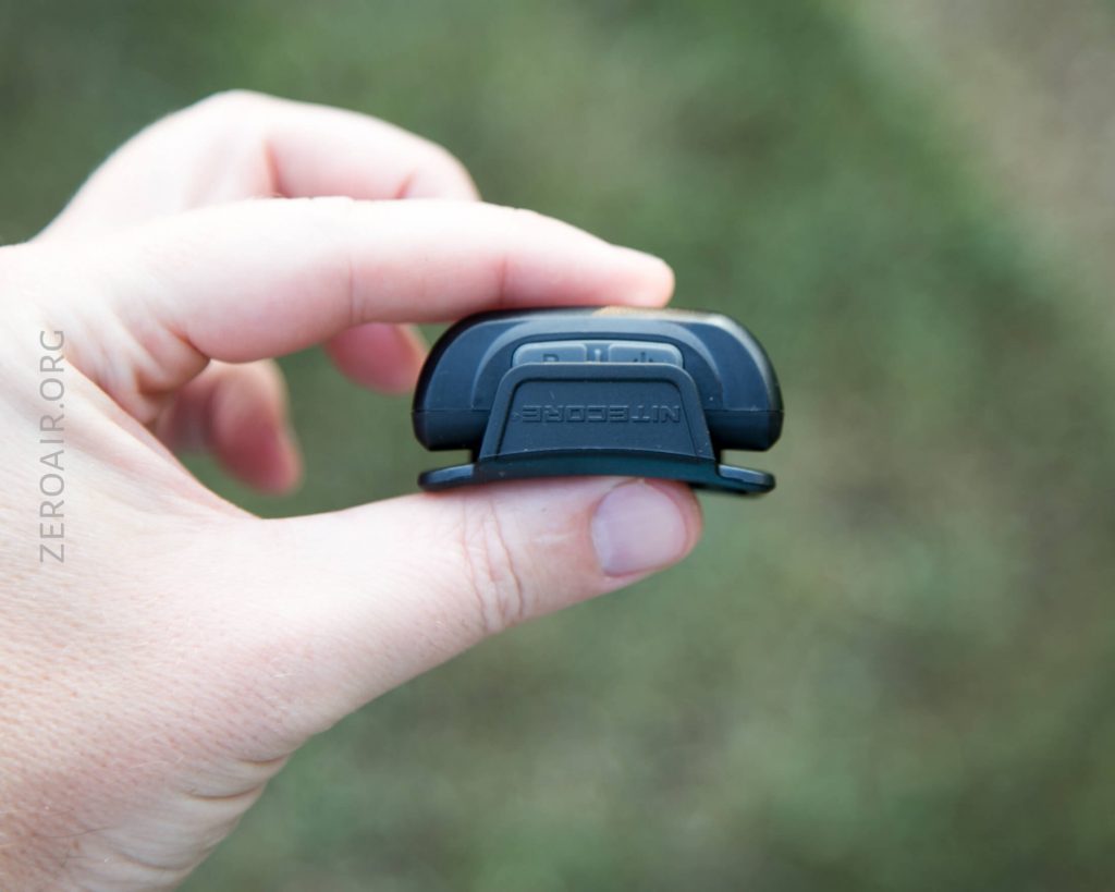 A hand holding a small black Nitecore device outdoors, with text NITECORE visible on the device and ZEROAIR.ORG watermarked on the image. The background is blurred grass.
