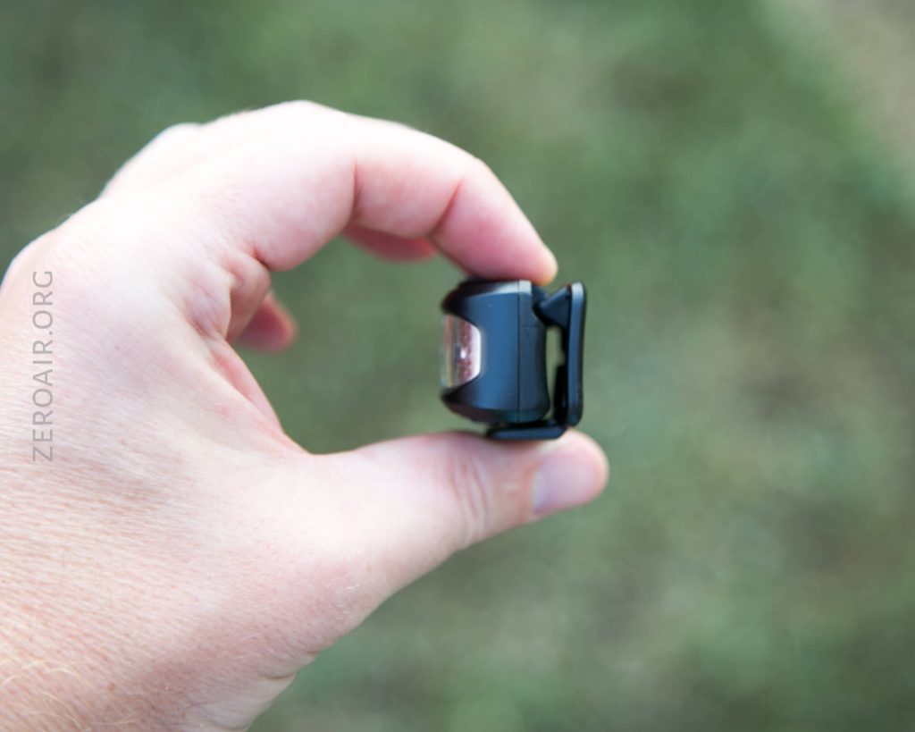 A hand holding a small black device with a clip, shown against a blurred green background. The device appears to be compact and rectangular, with a clear or reflective section in the center.