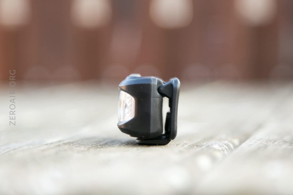 A small black bike light with a clear lens sits on a wooden surface outdoors, photographed in soft focus with a blurred background.