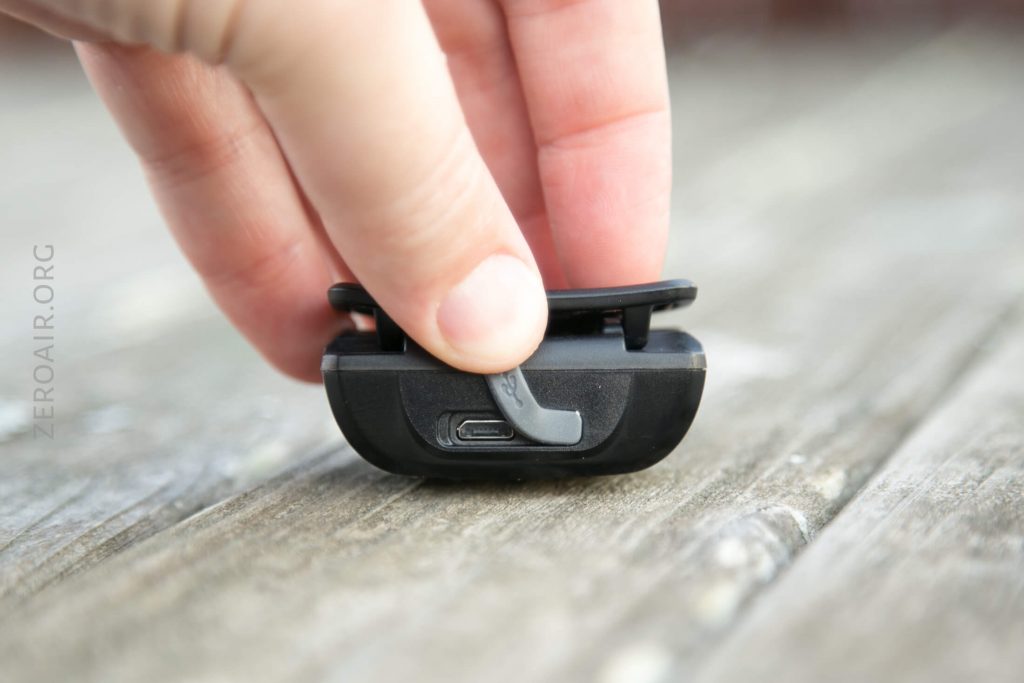 A hand lifts a rubber cover on a small black electronic device, revealing a micro USB port. The device is resting on a weathered wooden surface.