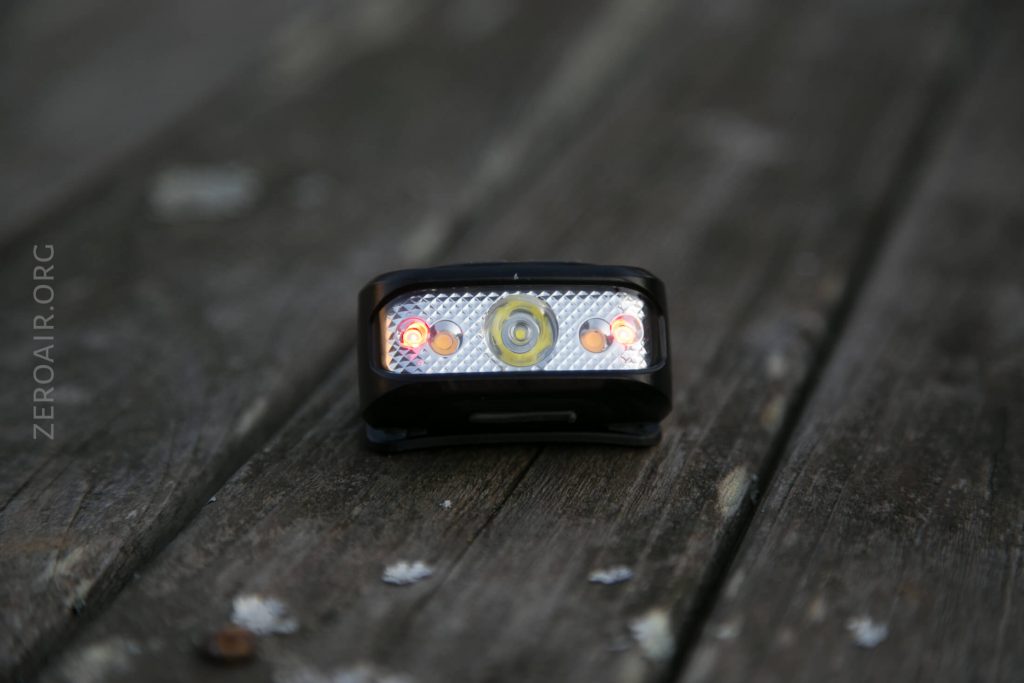 A small black LED headlamp with three illuminated lights sits on a weathered wooden surface. The background is out of focus, and the lamp face displays red and white LEDs. The text “ZEROAIR.ORG” is visible on the left.